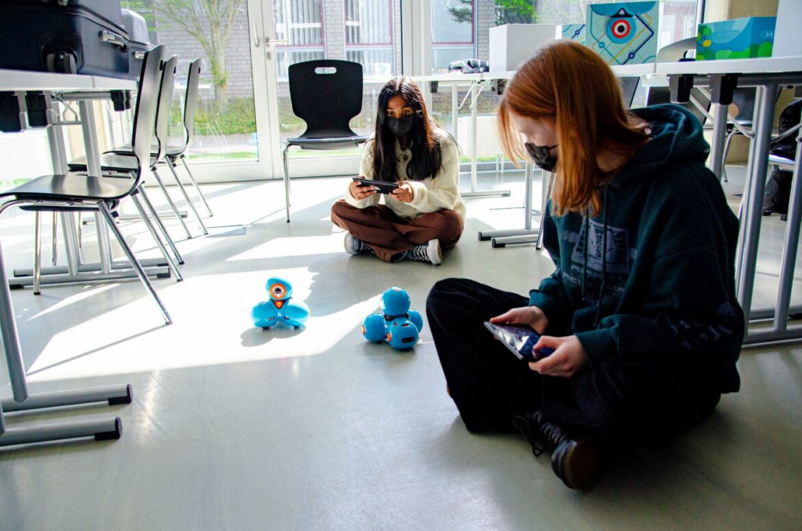 2022-fh-elektrotechnik Zwei Mädchen sitzen auf dem Boden und steuern zwei kleine Roboter, die umherfahren.