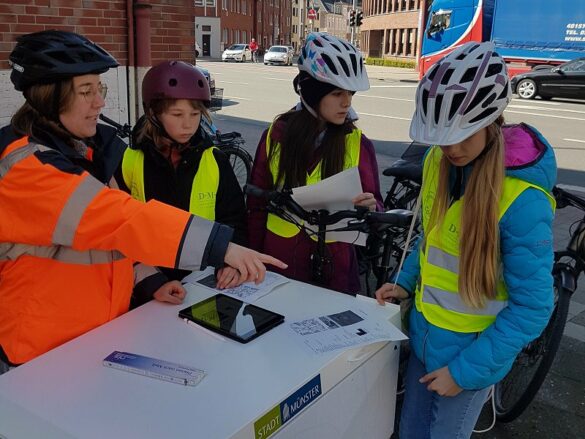 fahrradbuero-gd-2023-planungsgespraech-3 Besprechung der Fahrradplanerin mit den drei Schülerinnen an einer viel befahrenen Kreuzung.