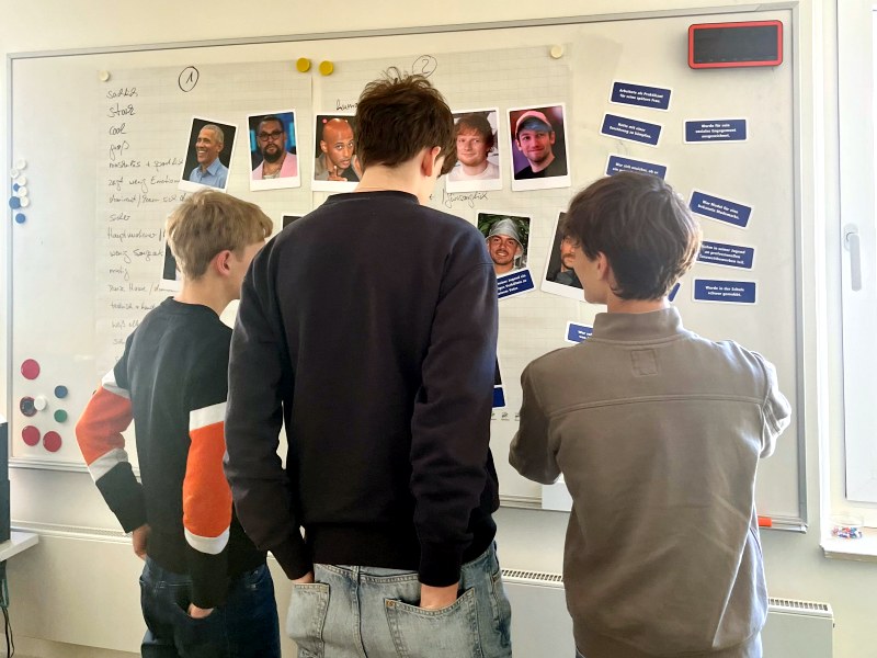 BoysDay-amt-17-2026 Drei Jungen stehen vor einer Moderationswand und diskutieren verschiedene Männlichkeitstypen, die als Fotos aufgehängt sind.