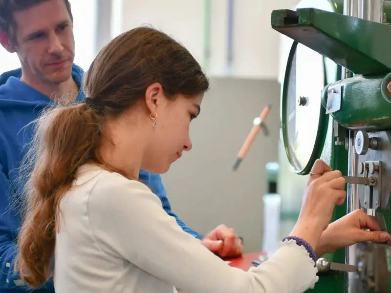 FH4-2026 Eine Schülerin arbeitet an einer Maschine, während ihr ein Anleiter über die Schulter schaut.